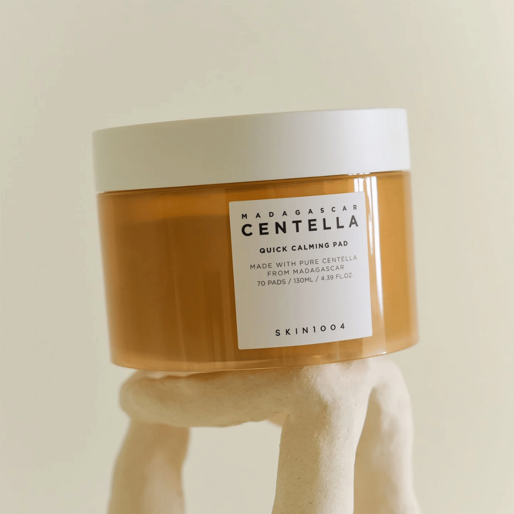 Słoik z SKIN1004 Madagascar Centella Quick Calming Pad umieszczony na rzeźbiarskiej białej piedestale – bursztynowy pojemnik z białą etykietą i pokrywką.