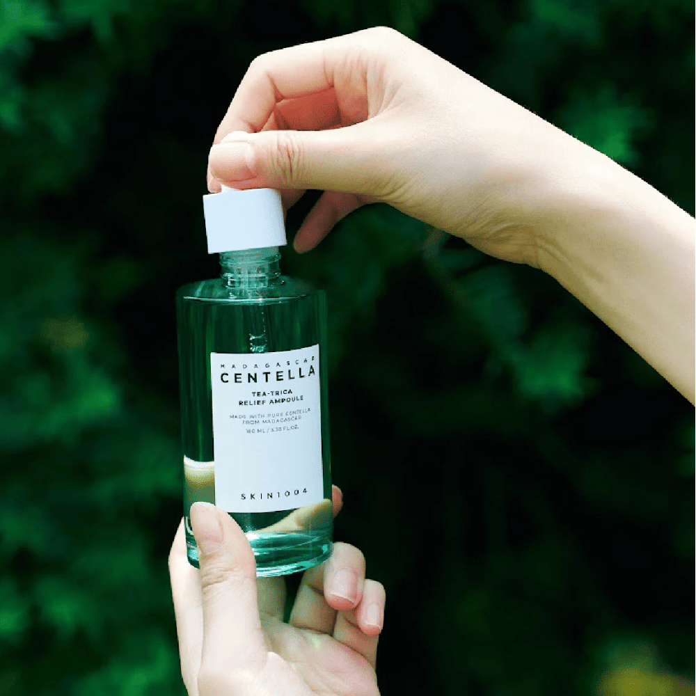 Osoba otwiera SKIN1004 Madagascar Centella Tea-Trica Relief Ampoule – zielona szklana butelka z białą etykietą trzymana w rękach na tle zieleni.