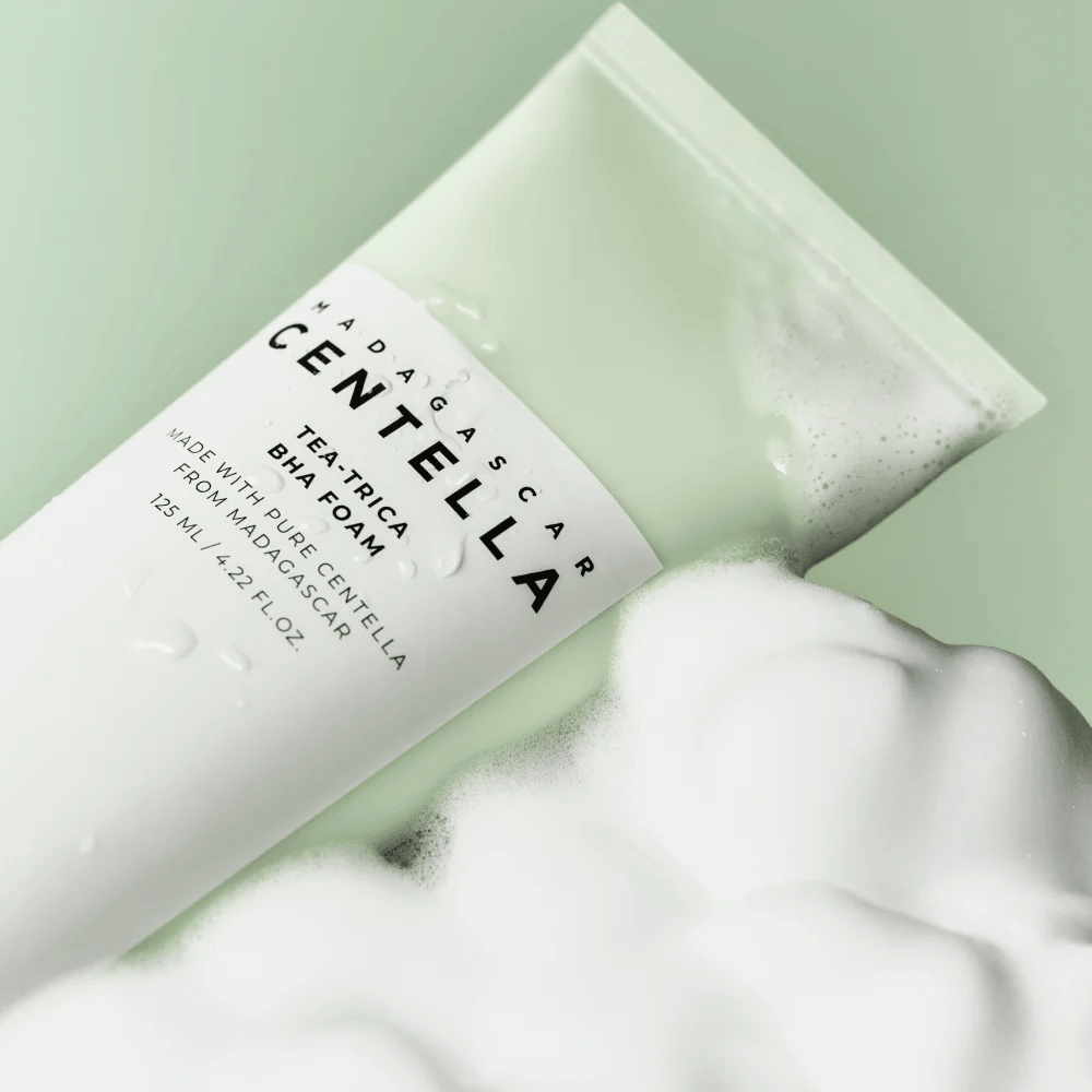 Produkt typu pianka oczyszczająca, "Madagascar Centella Tea-trica BHA Foam", jest częściowo widoczny z pianą wokół. Produkt ma zielono-biały design, a tekst "Madagascar Centella" i "Tea-trica BHA Foam" są wyraźnie widoczne. Objętość wynosi 125 ml.