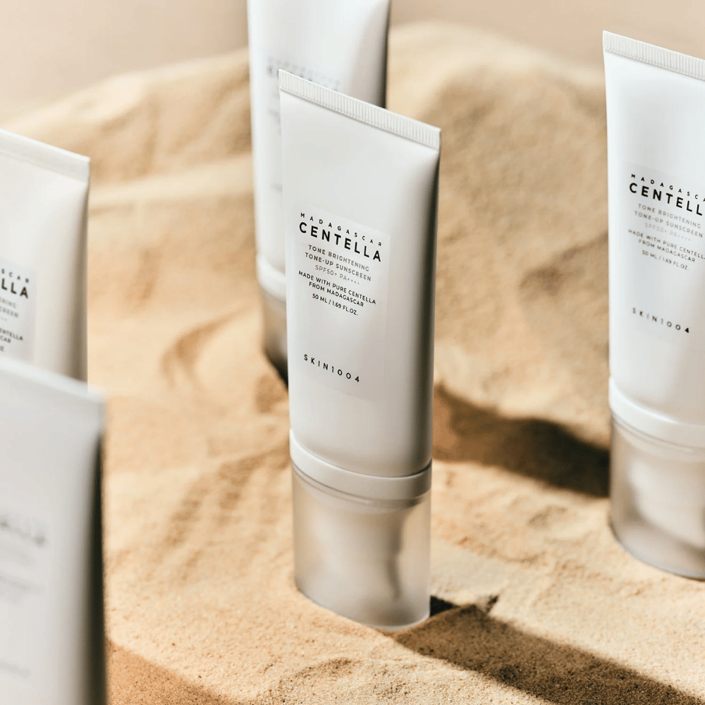Kilka tub z Madagascar Centella Tone Brightening Tone-Up Sunscreen stoi w piasku. Tuba jest biała z czarnym tekstem i zawiera 50 ml. Produkt ma SPF 50+ PA++++ i jest wykonany z czystej Centelli z Madagaskaru.