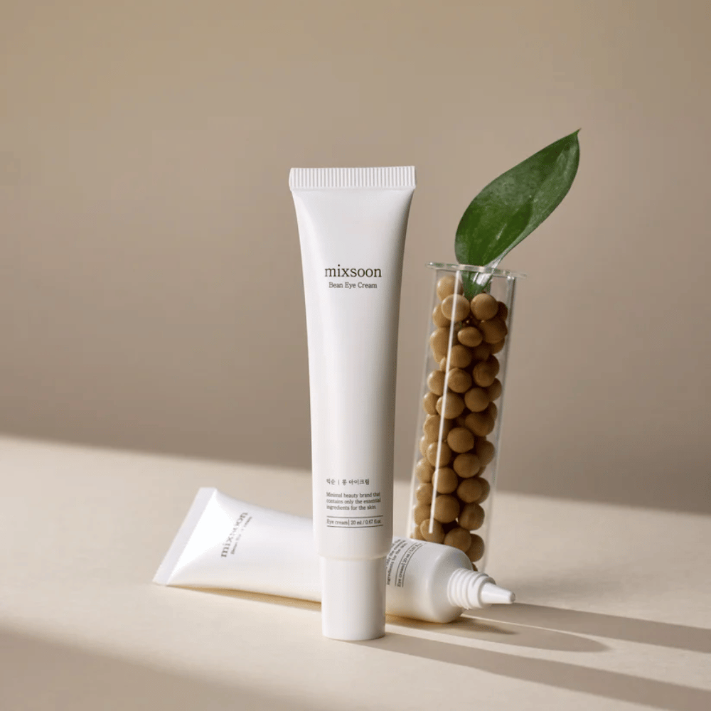 Dwie tubki Mixsoon Bean Eye Cream umieszczone obok probówki wypełnionej soją i zielonym liściem rośliny, na beżowej powierzchni z grą cieni.