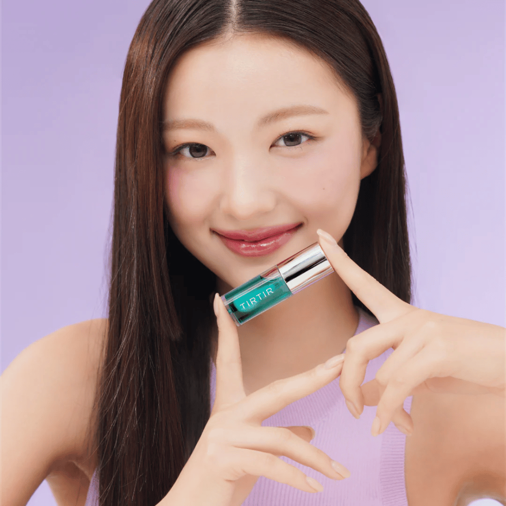 Kobieta z długimi, prostymi włosami trzyma TIRTIR My Glow Lip Oil i prezentuje swoje błyszczące, różowe usta na fioletowym tle.
