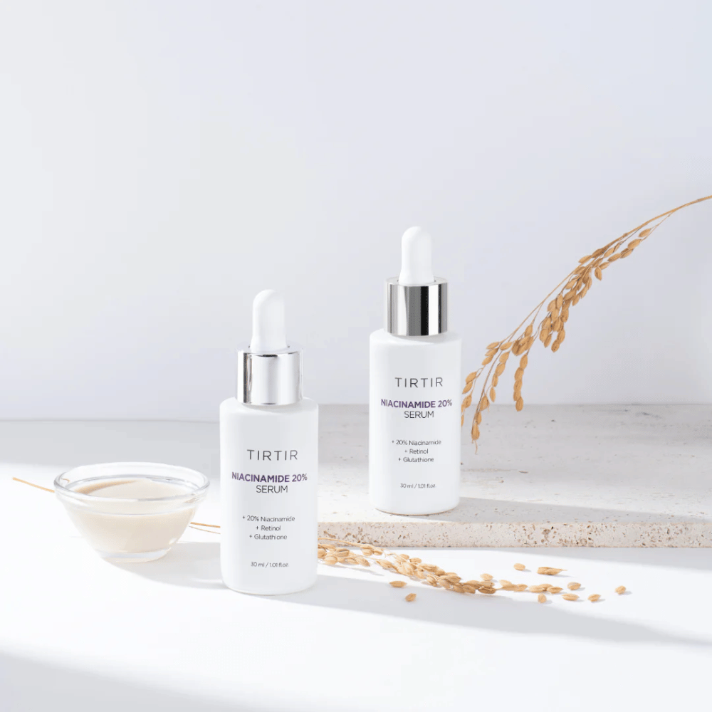 Dwie butelki Tirtirs "Niacinamide 20% Serum" są pokazane na jasnej powierzchni wraz z małą miseczką z kremową substancją i kilkoma źdźbłami suchych roślin. Serum zawiera 20% niacynamidu, retinolu i glutationu, co przyczynia się do poprawy wyglądu i tekstury skóry.