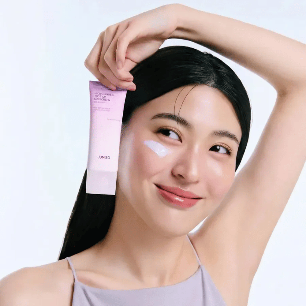 Modelka trzyma JUMISO Niacinamide 3+ Tone-Up Sunscreen SPF50+ PA++++ – krem przeciwsłoneczny z niacynamidem, który wyrównuje koloryt skóry i chroni skórę.