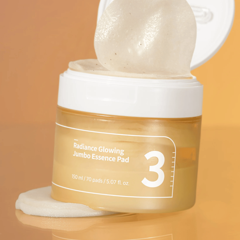 Otwarty słoik z numbuzin No. 3 Radiance Glowing Jumbo Essence Pad, jeden pad widoczny oparty o krawędź, tło w pomarańczowym tonie.