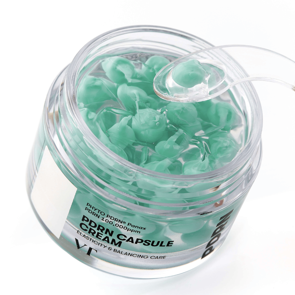 Zbliżenie na otwarty słoik z VT PDRN Capsule Cream. Zawartość składa się z małych zielonych kapsułek w przezroczystym żelu, z dołączoną szpatułką do aplikacji. Tekst na słoiku brzmi "Elasticity & Balancing Care".