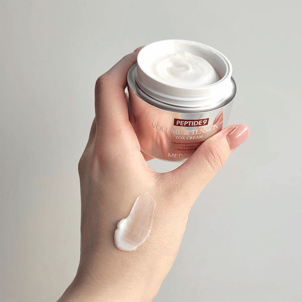 Ręka trzyma MEDI-PEEL Peptide 9 Volume & Tension Tox Cream z odrobiną kremu na grzbiecie dłoni. Jedwabista formuła nawilża i nadaje skórze jędrność i sprężystość.