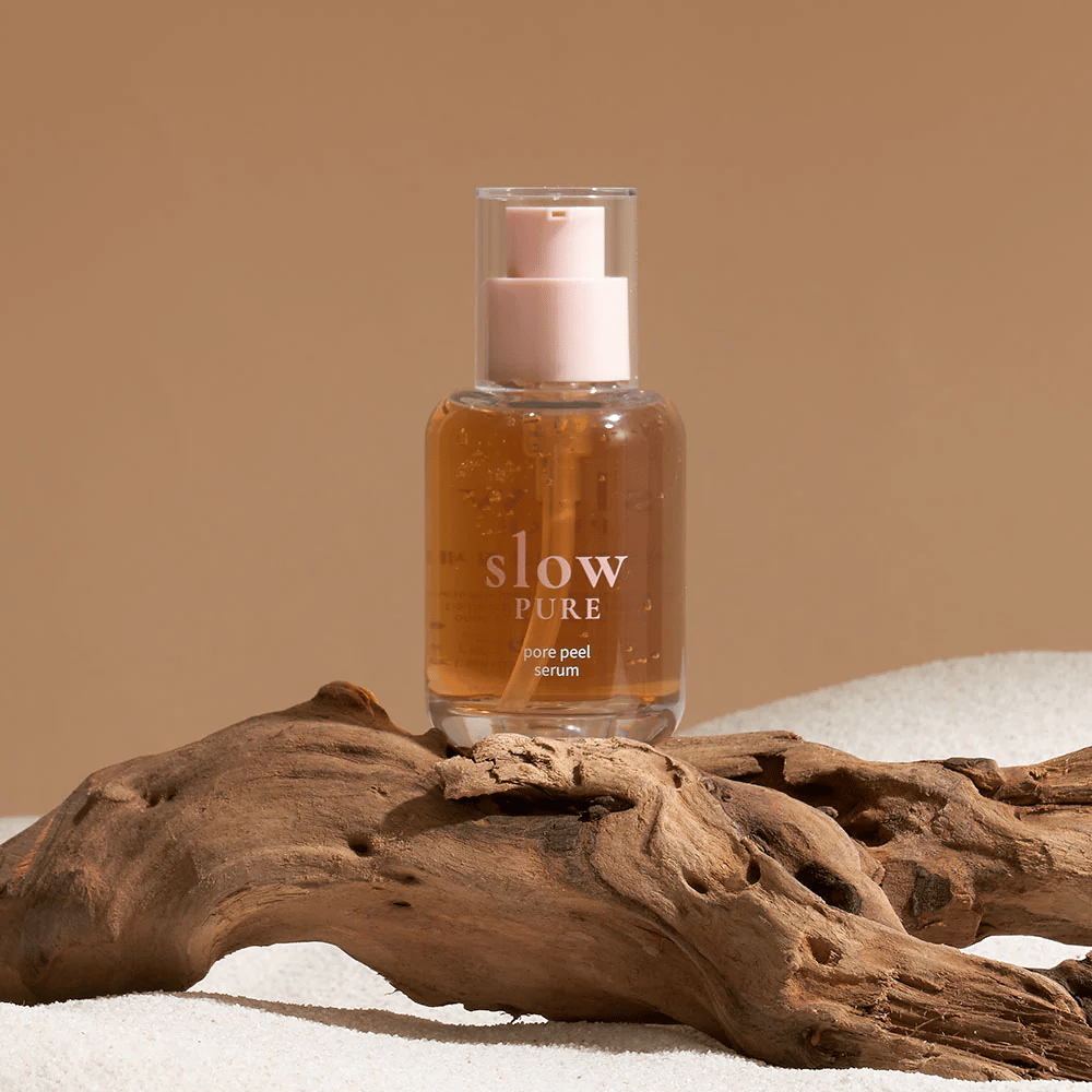 Slow Pure Pore Peel Serum umieszczony na piasku z drewnianym detalem w tle, symbol naturalnej pielęgnacji skóry.