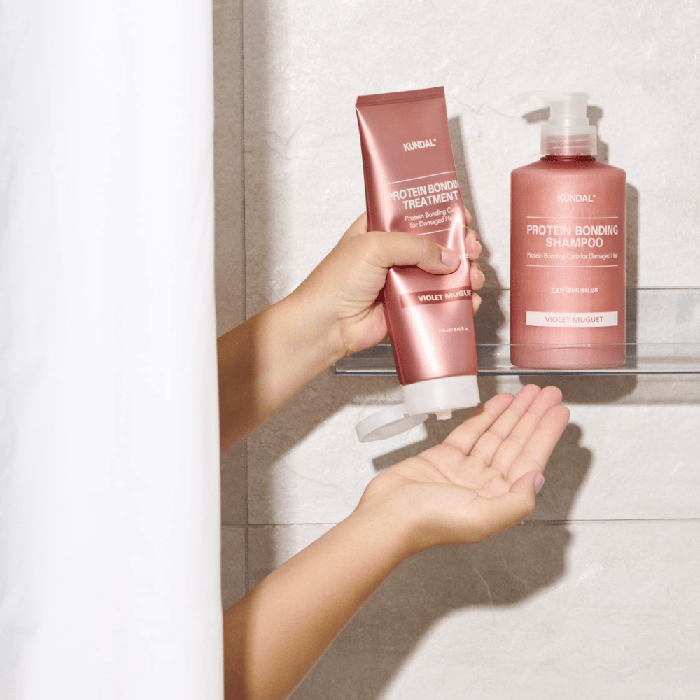 KUNDAL Protein Bonding Treatment Violet Muguet używane jest pod prysznicem, z Protein Bonding Shampoo umieszczonym na półce obok.