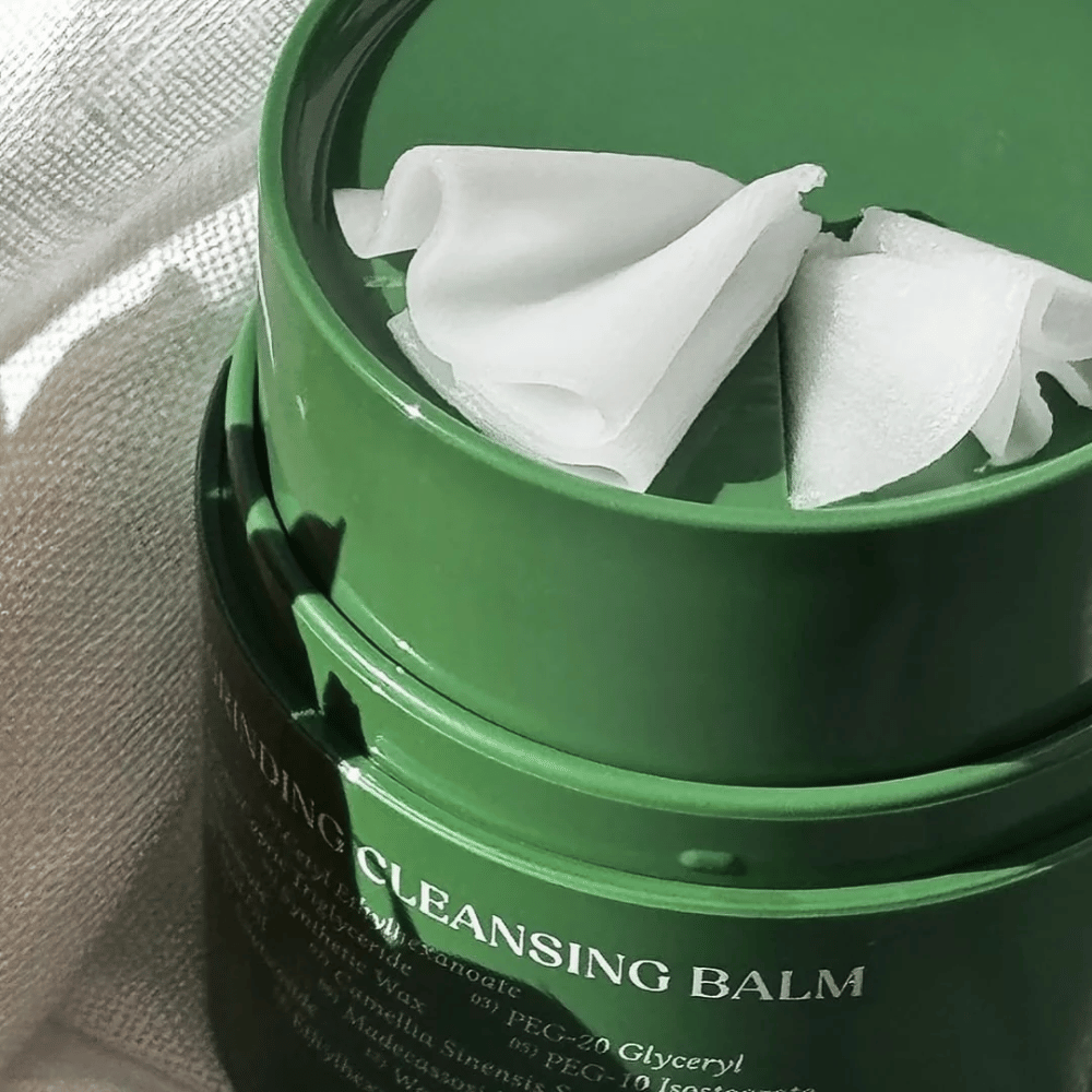 Obraz przedstawia zielony słoik z "Cleansing Balm", który jest otwarty, aby ukazać dołączoną szpatułkę i miękką zawartość. Kremowa konsystencja odbija światło, co daje poczucie luksusu i skuteczności w oczyszczaniu.