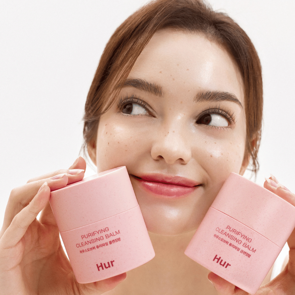 Model trzyma dwa słoiki Jak Purifying Cleansing Balm. Łagodny balsam oczyszczający, który nadaje się do wrażliwej skóry i daje świeże, nawilżone uczucie po oczyszczeniu.