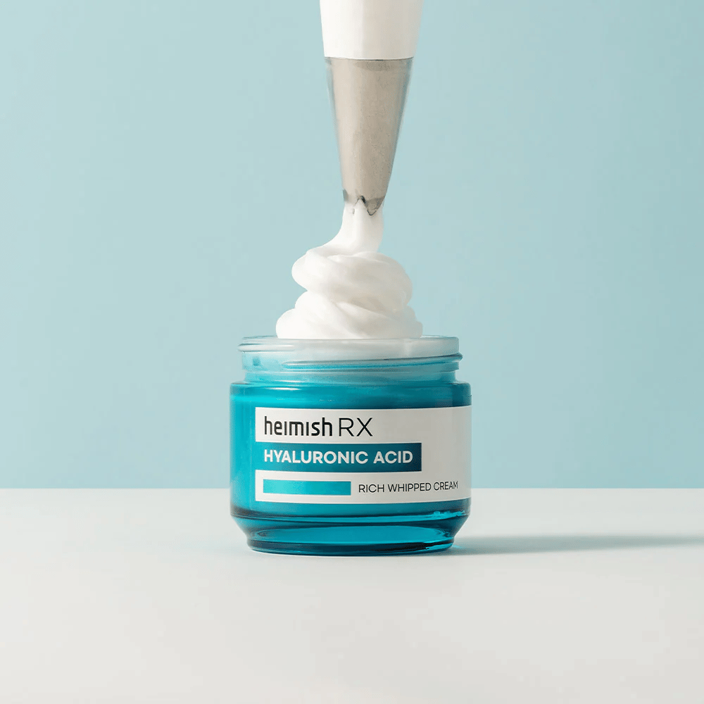 Heimish RX Hyaluronic Acid Rich Whipped Cream w niebieskim szklanym słoiku, gdzie worek do wyciskania nakłada puszysty krem. Nawilżający krem do twarzy z kwasem hialuronowym dla intensywnego nawilżenia i miękkiej, pełnej skóry.