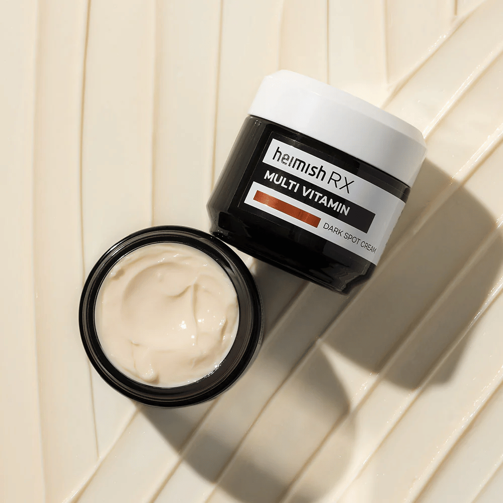 heimish RX Multi Vitamin Dark Spot Cream – odżywczy krem do twarzy do redukcji ciemnych plam i nadania skórze bardziej jednolitego kolorytu. Jedwabista konsystencja kremu prezentowana jest w otwartym słoiku z eleganckim opakowaniem.