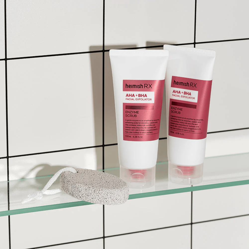 Dwie tubki Heimish RX AHA + BHA Facial Exfoliator umieszczone na szklanej półce w łazience z białą ścianą z płytek. Obok produktów leży pumeks z białym sznurkiem. Stylowe i minimalistyczne zdjęcie pielęgnacji skóry.