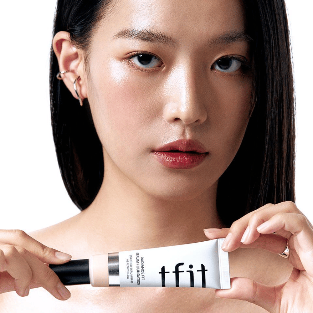 Modelka o błyszczącej, gładkiej skórze trzyma TFIT Radiance Fit Serum Foundation, co podkreśla naturalne wykończenie i blask produktu.