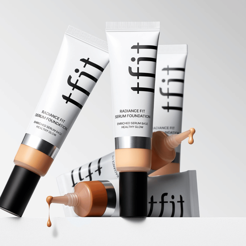 Kilka tubek TFIT Radiance Fit Serum Foundation w różnych odcieniach, z których jedna z foundation kapie na jasnoszare tło.