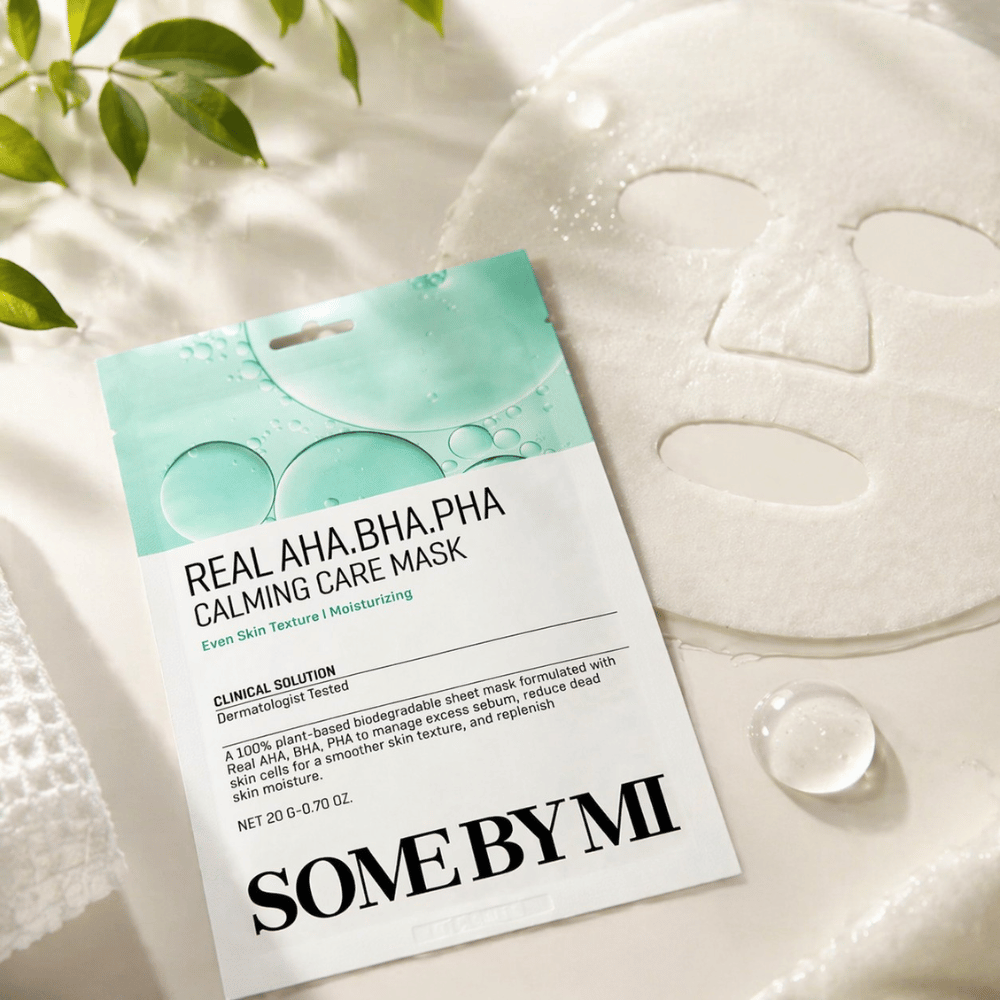 SOME BY MI Real AHA.BHA.PHA Calming Care Mask jest pokazana wraz z rozwiniętą maską w płachcie. Wokół produktu widoczne są krople wilgoci i zielone liście, które wzmacniają wrażenie nawilżenia, kojącej pielęgnacji i delikatnego złuszczania. Obraz podkreśla nacisk maski na bardziej równomierną teksturę skóry i poprawioną równowagę nawilżenia.