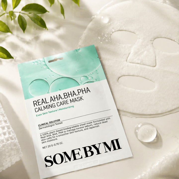 SOME BY MI Real AHA.BHA.PHA Calming Care Mask jest pokazana wraz z rozwiniętą maską w płachcie. Wokół produktu widoczne są krople wilgoci i zielone liście, które wzmacniają wrażenie nawilżenia, kojącej pielęgnacji i delikatnego złuszczania. Obraz podkreśla nacisk maski na bardziej równomierną teksturę skóry i poprawioną równowagę nawilżenia.