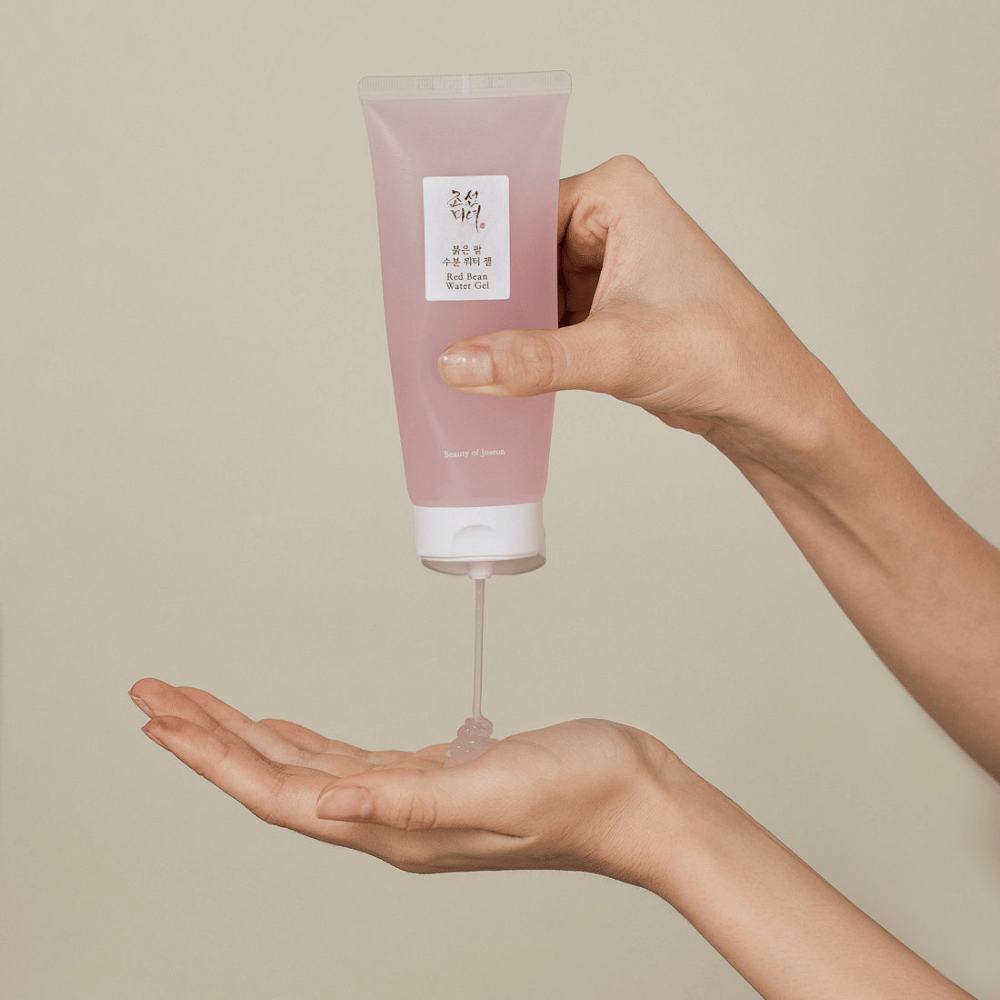 Obraz pokazuje osobę trzymającą tubkę "Red Bean Water Gel" z Beauty of Joseon i wyciskającą przezroczysty żel na dłoń. Produkt wydaje się być przeznaczony do pielęgnacji skóry i ma atrakcyjne, delikatnie różowe opakowanie.