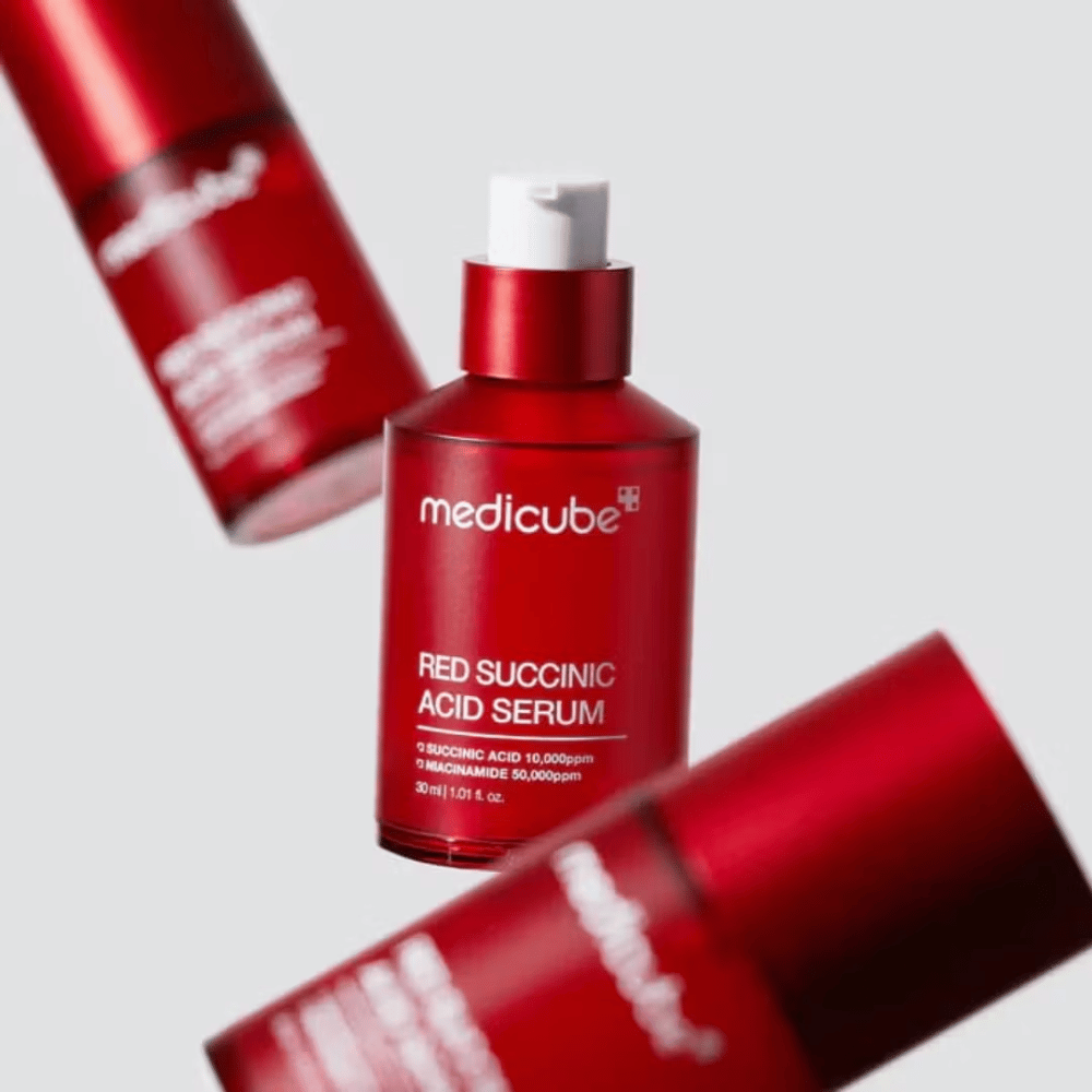 Medicube Red Succinic Acid Serum w czerwonej butelce z pompką, otoczony przez więcej identycznych butelek na jasnym tle. Produkt zawiera kwas bursztynowy i niacynamid do leczenia trądziku i plam pigmentacyjnych.