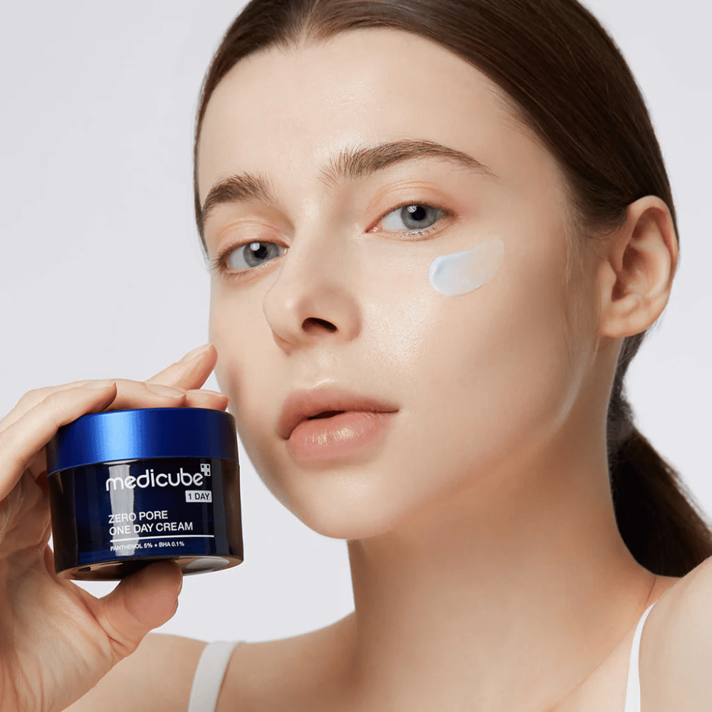 Kobieta z naturalnym wyglądem trzyma Medicube Zero Pore One Day Cream. Niewielka ilość kremu jest nałożona na jej policzek. Produkt jest zaprojektowany, aby poprawić strukturę skóry i zminimalizować wielkość porów, z lekką i nawilżającą formułą.