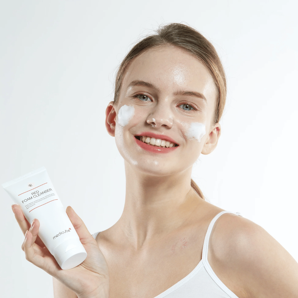 Młoda kobieta z pianką oczyszczającą na twarzy trzyma tubkę Medicube Red Foam Cleanser i uśmiecha się do kamery. Ma na sobie białą koszulkę na ramiączkach, a tło jest jasne i proste.