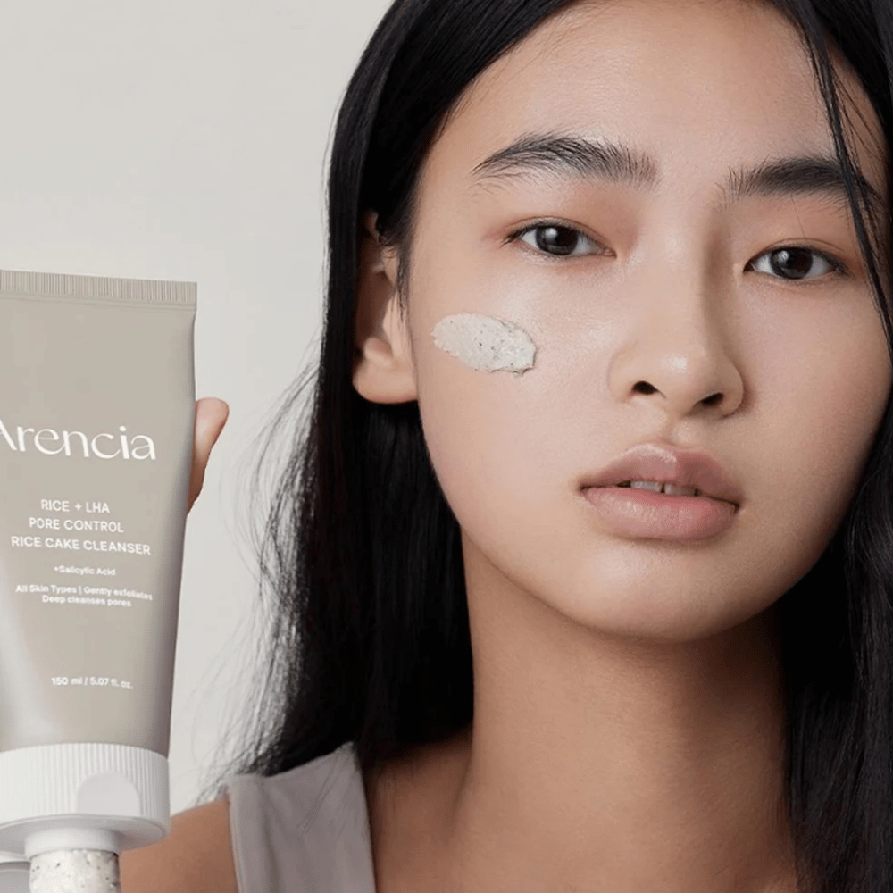 Model trzymający Arencia Rice + LHA Pore Control Rice Cake Cleanser z nałożonym produktem na policzku, ilustruje łagodne, ale skuteczne oczyszczanie porów do codziennego użytku.