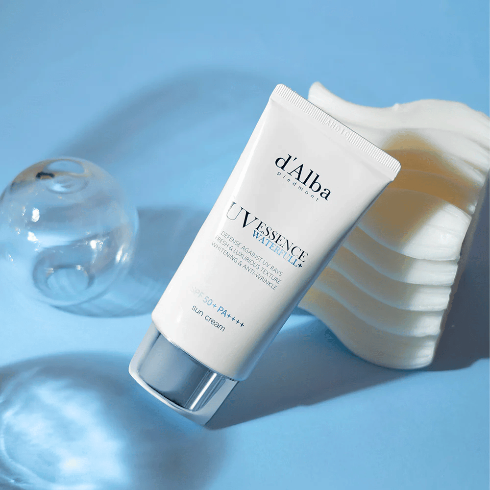 d'Alba UV Essence Waterfull+ Sun Cream z SPF 50+ PA++++ leży na niebieskim tle z odbiciami światła i dekoracyjnymi elementami – podkreśla świeże i nawilżające właściwości kremu przeciwsłonecznego.