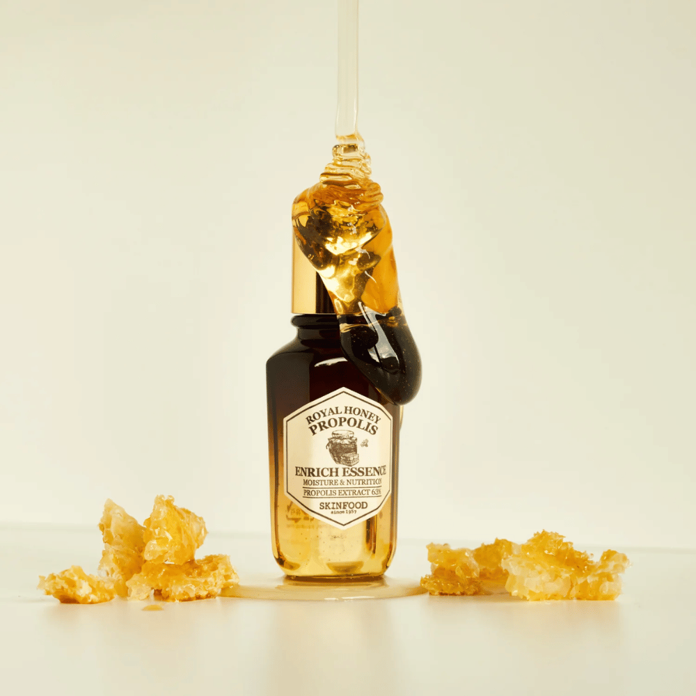 Obraz szklanej butelki z Royal Honey Propolis Enrich Essence od Skinfood. Duża kropla złotego miodu spływa z złotego wieczka butelki. Wokół butelki leżą małe kawałki plastra miodu rozrzucone na jasnej powierzchni. Esencja jest reklamowana z naciskiem na wilgoć i odżywianie, z 63% ekstraktem z propolisu.