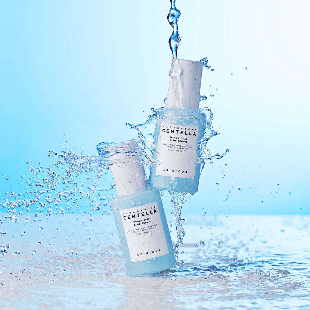 Dwie butelki SKIN1004 Madagascar Centella Hyalu-Cica Blue Serum uderzone przez kaskadę wody na jasnoniebieskim tle.