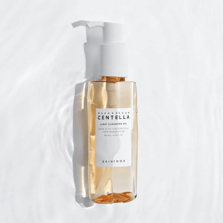 SKIN1004 Madagascar Centella Light Cleansing Oil på vit bakgrund med mjuka vattenreflexer som framhäver den transparenta flaskan och gyllene innehållet.