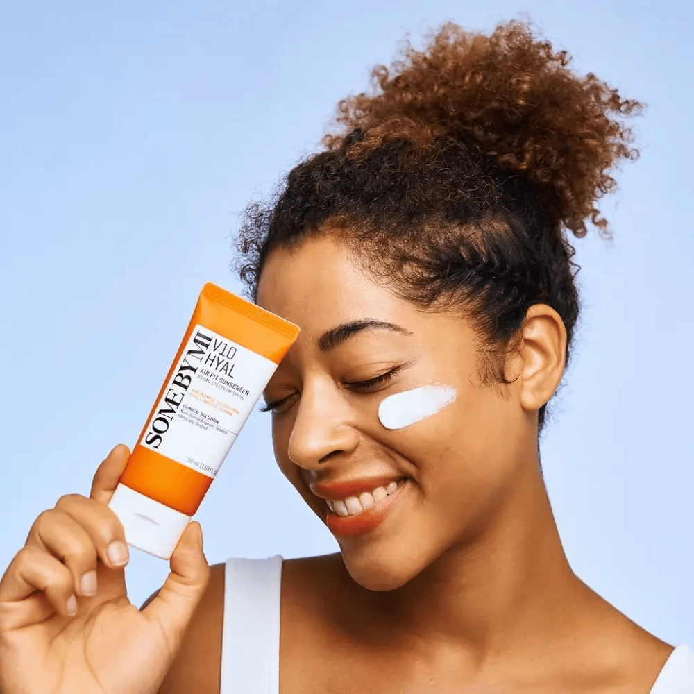 Kobieta trzyma tubkę z SOME BY MI V10 Hyal Air Fit Sunscreen i ma białą plamę kremu przeciwsłonecznego na policzku. Uśmiecha się szeroko i wyraźnie pokazuje produkt, co podkreśla jego zastosowanie i korzyści dla pielęgnacji skóry oraz ochrony przed słońcem.