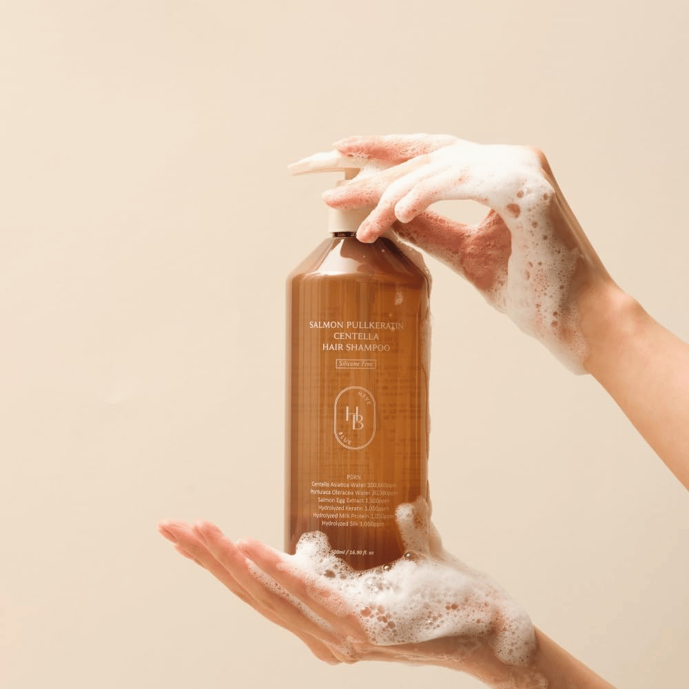 Heveblue Salmon Pullkeratin Centella Hair Shampoo w brązowej butelce otoczonej pianą. Produkt jest trzymany między dwiema dłońmi i podkreśla oczyszczające oraz pielęgnacyjne działanie pielęgnacji włosów z obfitą pianą.