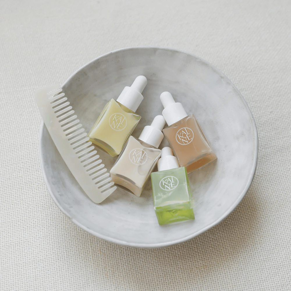 Cztery małe butelki z serum od KAINÉ Serum Discovery Kit leżą w białej misce obok grzebienia, na tle naturalnym i spokojnym w beżowym tonie.