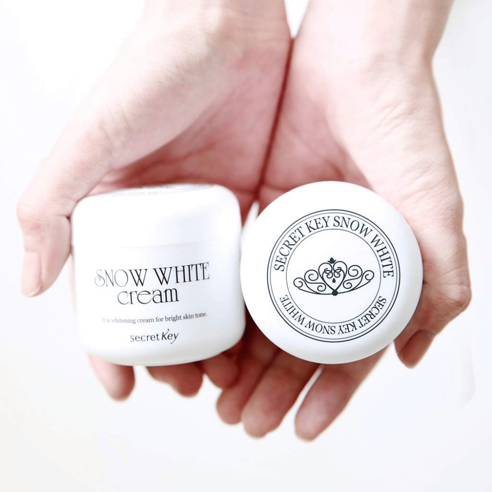 Två burkar Secret Key Snow White Cream hålls i händerna, en visar locket och en visar framsidan.