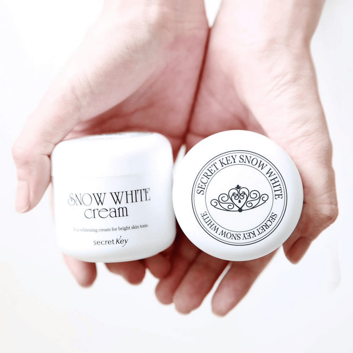 Två burkar Secret Key Snow White Cream hålls i händerna, en visar locket och en visar framsidan.