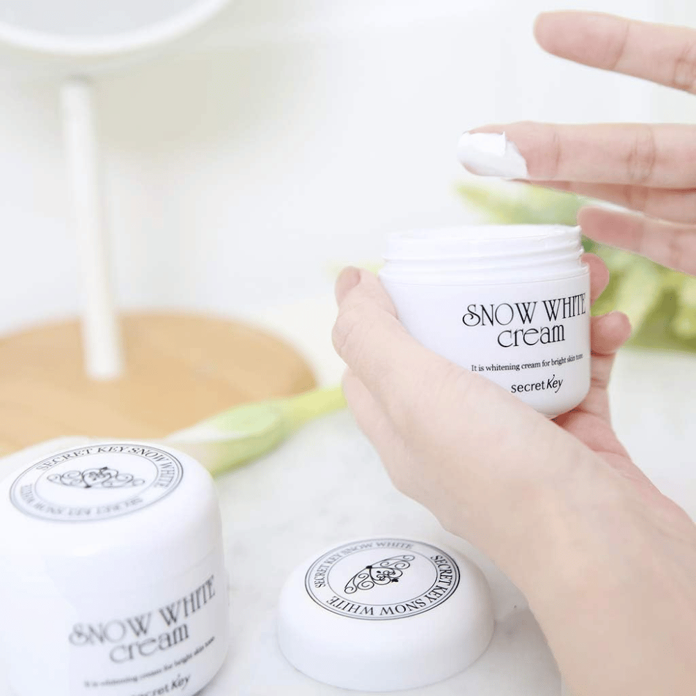 Ręka wyciągająca biały krem ze słoika Secret Key Snow White Cream na toaletce.