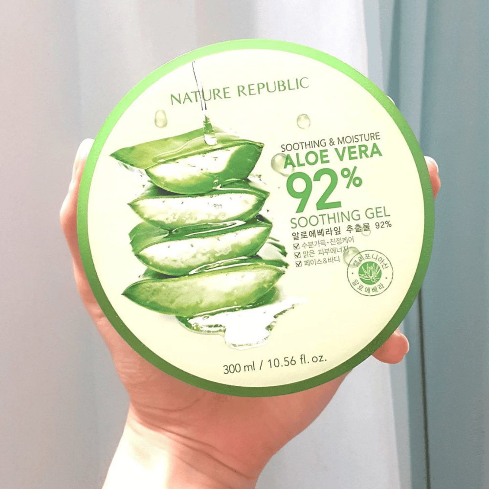 Ręka trzyma okrągłą puszkę z Nature Republic Aloe Vera 92% Soothing Gel. Puszka jest zielona z obrazem aloesu i tekstem opisującym jej kojące i nawilżające właściwości. Zawartość to 300 ml.