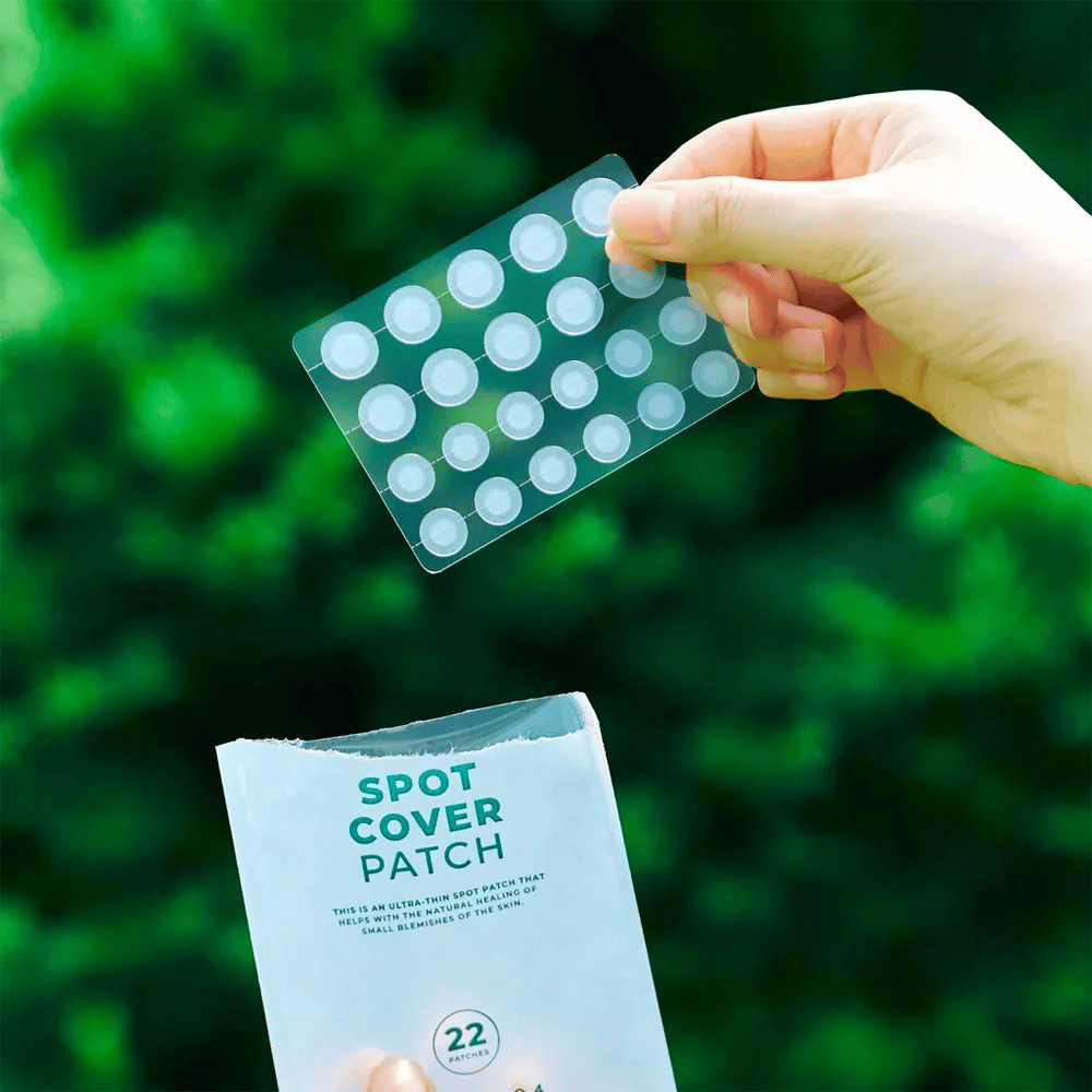 Ręka trzyma arkusz z łatkami Spot Cover, które są przezroczyste i zaokrąglone. W tle widać rozmyte zielone otoczenie. Opakowanie pokazuje część opakowania z tekstem "SPOT COVER PATCH" oraz informację, że produkt zawiera 22 plastry, które pomagają w leczeniu małych obszarów skóry.