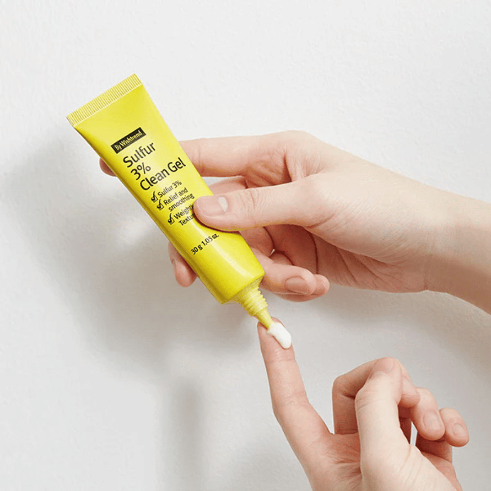 Zbliżenie na ręce aplikujące By Wishtrend Sulfur 3% Clean Gel, żółtą tubkę z białym żelem, na białym tle.