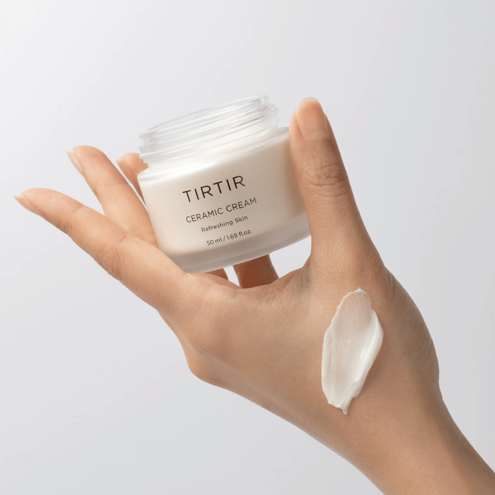 Ręka trzyma słoik z TIRTIR Ceramic Cream. Na grzbiecie ręki nałożono ilość kremu. Krem wydaje się być gęsty i nawilżający. Opakowanie wskazuje, że zawartość jest przeznaczona do "Orzeźwiającej Skóry" i ilość wynosi 50 ml / 1.69 fl oz.