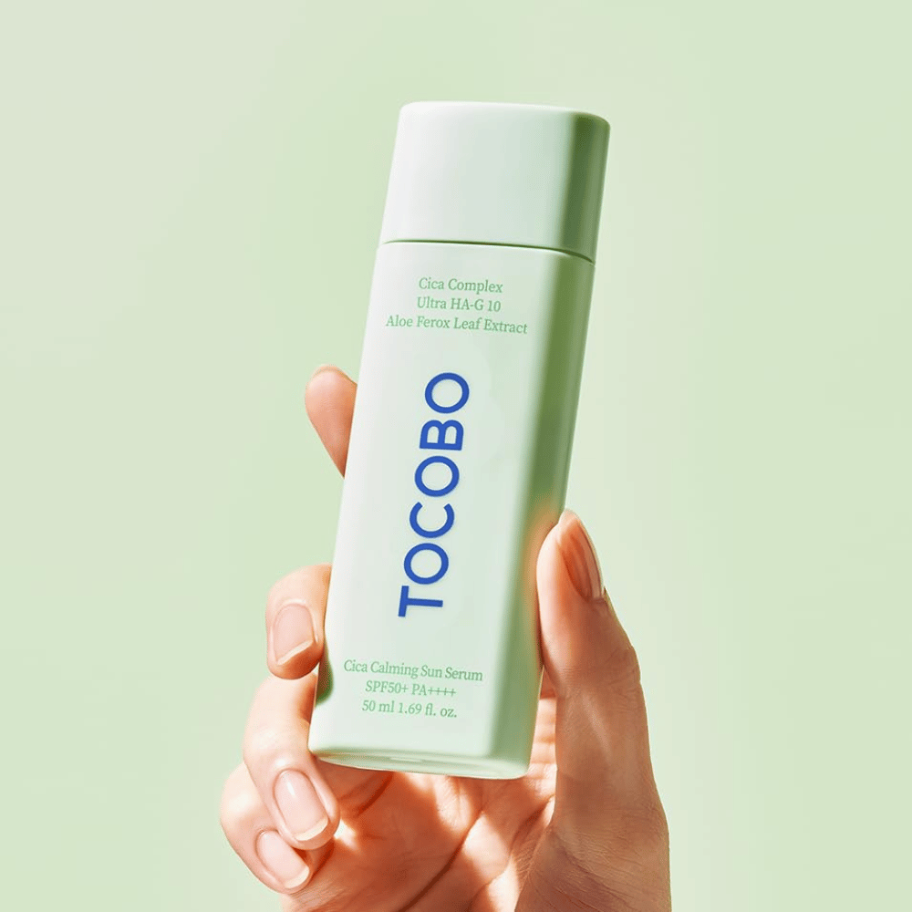Ręka trzyma TOCOBO Cica Calming Sun Serum w miętowozielonym opakowaniu z niebieskim napisem na jasnozielonym tle.