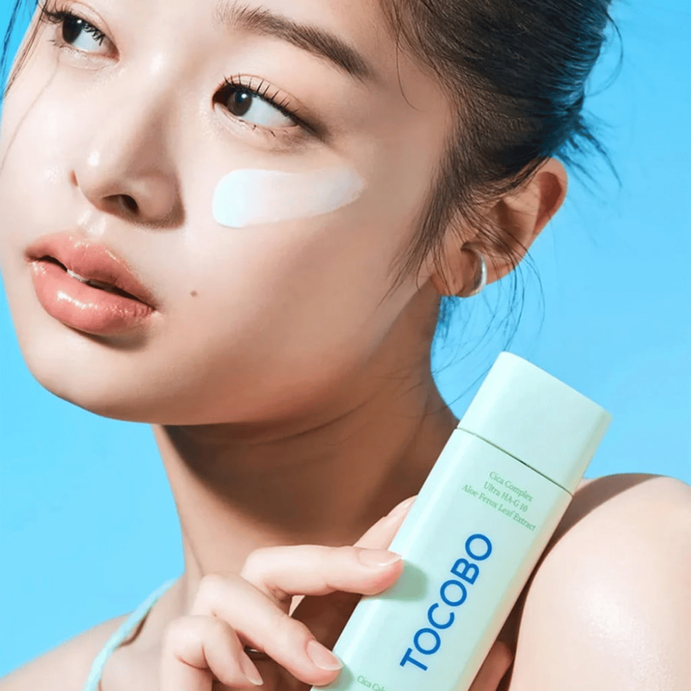 Modelka z kremem przeciwsłonecznym pod okiem trzyma TOCOBO Cica Calming Sun Serum w miętowozielonym opakowaniu na niebieskim tle.