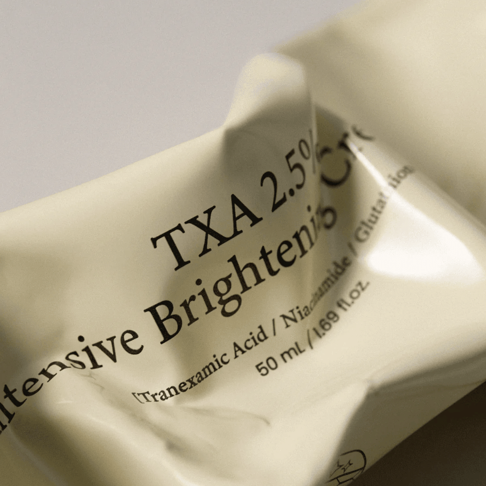 Zbliżenie na AXIS - Y TXA 2.5% Intensive Brightening Cream, gdzie miękki i minimalistyczny design tubki jest w centrum uwagi. Zdjęcie podkreśla nazwę produktu, Tranexamic Acid i Niacinamide oraz eleganckie beżowe opakowanie.