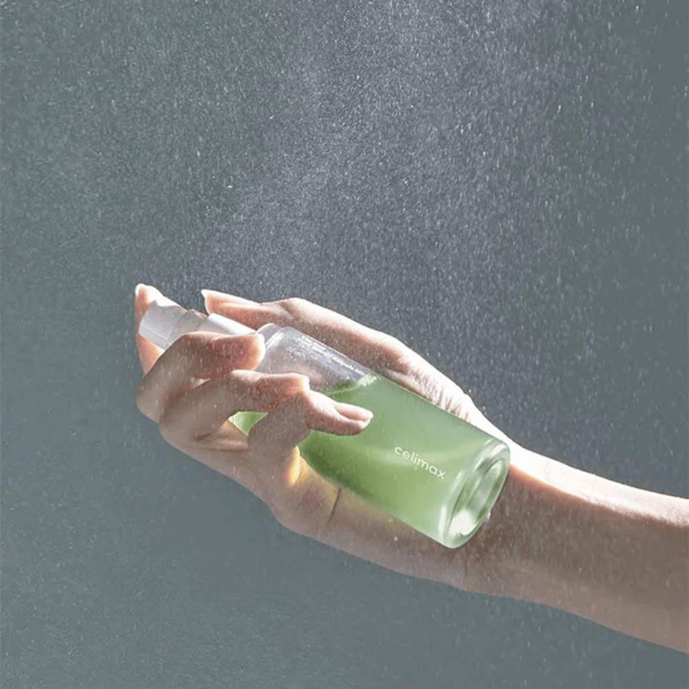 Ręka trzymająca Celimax spray Noni Mist i aplikująca produkt w delikatnej, równomiernej mgiełce na skórze. Butelka jest zielona, a produkt reklamowany jest jako "The Real Noni Energy Ampule Mist" do dostarczania energii i nawilżania skóry.