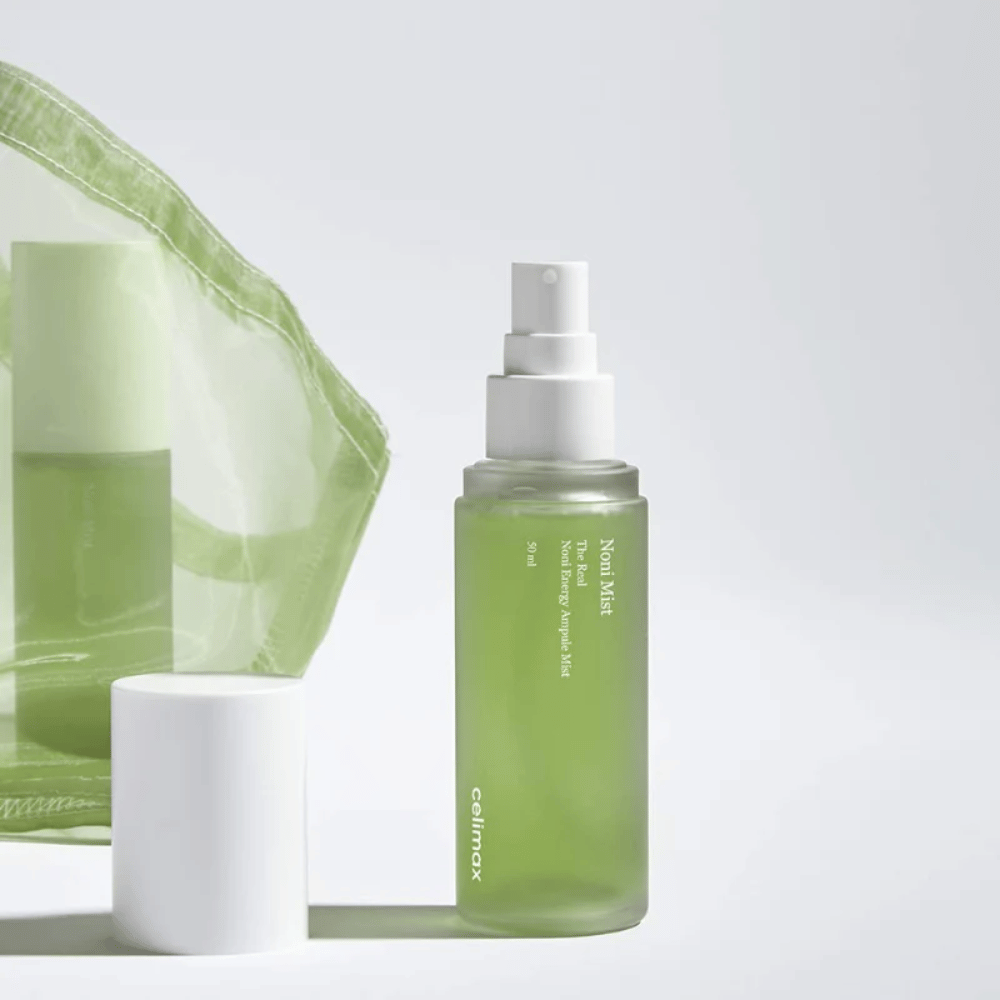 Butelka z Celimax Noni Mist, zielona butelka z białą pompką, stojąca obok nakrętki. Produkt jest zapakowany w przezroczystą plastikową torbę i reklamowany jako Noni Energy Ampule Mist.