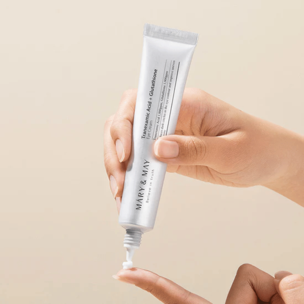 Ręka trzyma tubkę MARY & MAY "Tranexamic Acid + Glutathione Eye Cream". Tubka jest biała, a jej design jest czysty i prosty, co podkreśla kojące i rozjaśniające właściwości produktu dla okolic oczu. Produkt jest aplikowany delikatnie, z naciskiem na precyzję i skuteczność.