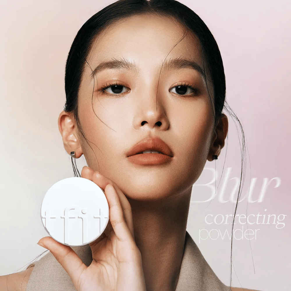 Kobieta z nieskazitelnym makijażem trzyma okrągłą białą puszkę z logo TFIT, z napisem "Blur Correcting Powder" w tle.