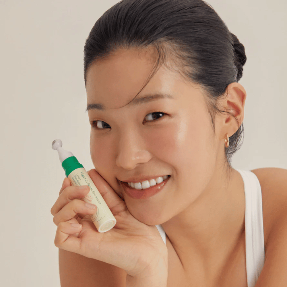 Kobieta z upiętymi włosami i złotymi kolczykami trzyma AXIS-Y Vegan Collagen Eye Serum 10ml blisko twarzy, podczas gdy delikatnie się uśmiecha. Serum jest zaprojektowane, aby nawilżać i redukować zmarszczki wokół oczu, specjalnie opracowane dla wrażliwej skóry.