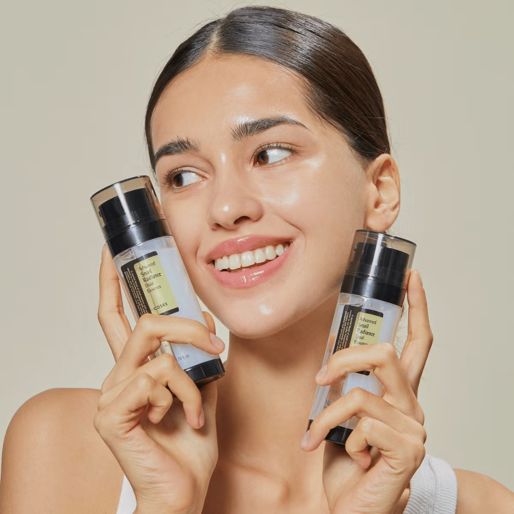 Uśmiechnięta kobieta z błyszczącą skórą trzyma dwie butelki COSRX Advanced Snail Radiance Dual Essence 80 ml blisko twarzy na jasnym tle.
